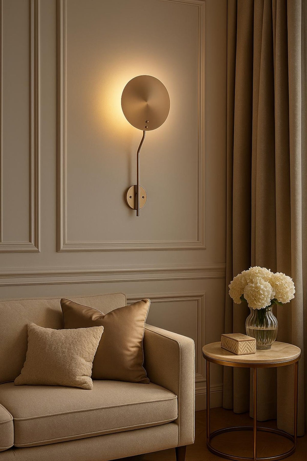 Brass disc wall sconce installed on paneled wall above sofa, creating soft ambient halo lighting in a warm neutral room.