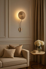 Brass disc wall sconce installed on paneled wall above sofa, creating soft ambient halo lighting in a warm neutral room.