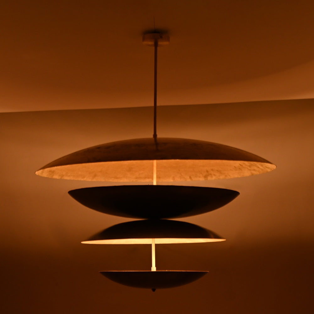 Modern black and gold chandelier illuminated in a warm ambient glow, featuring four dome-shaped tiers suspended on a central rod, casting layered light patterns on the ceiling and wall.