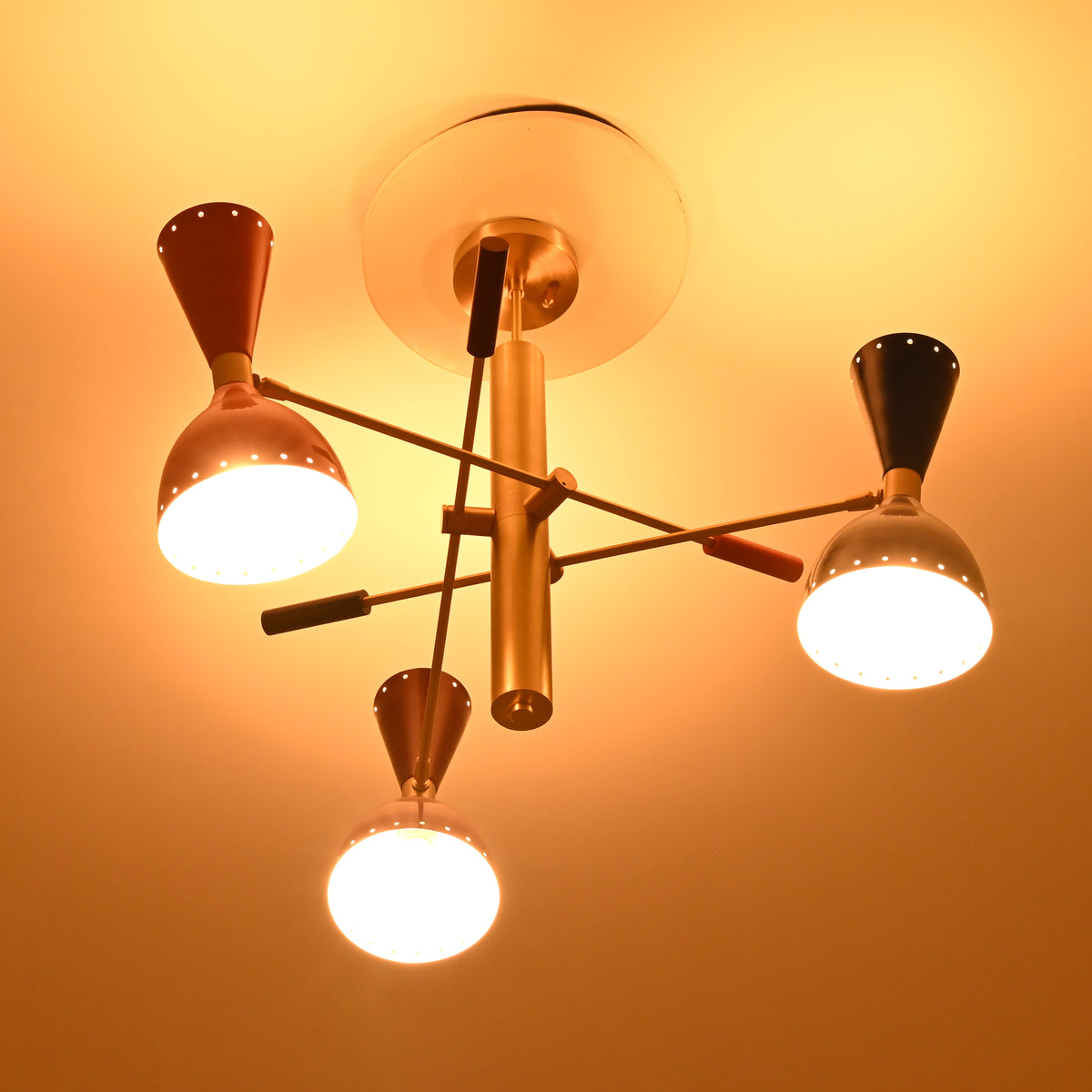 Angled night view of Manhattan retro chandelier showcasing asymmetric brass-tone arms, colorful metal shades, and bright downward illumination.