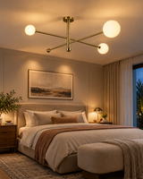 Marrakech brass linear globe chandelier illuminating an elegant contemporary bedroom with an upholstered bed, neutral bedding, wooden bedside tables, soft curtains, and warm ambient lighting.