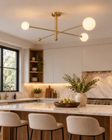 Marrakech brass linear globe chandelier suspended above a marble kitchen island with upholstered counter stools, white cabinetry, walnut accents, and natural daylight in a modern kitchen.