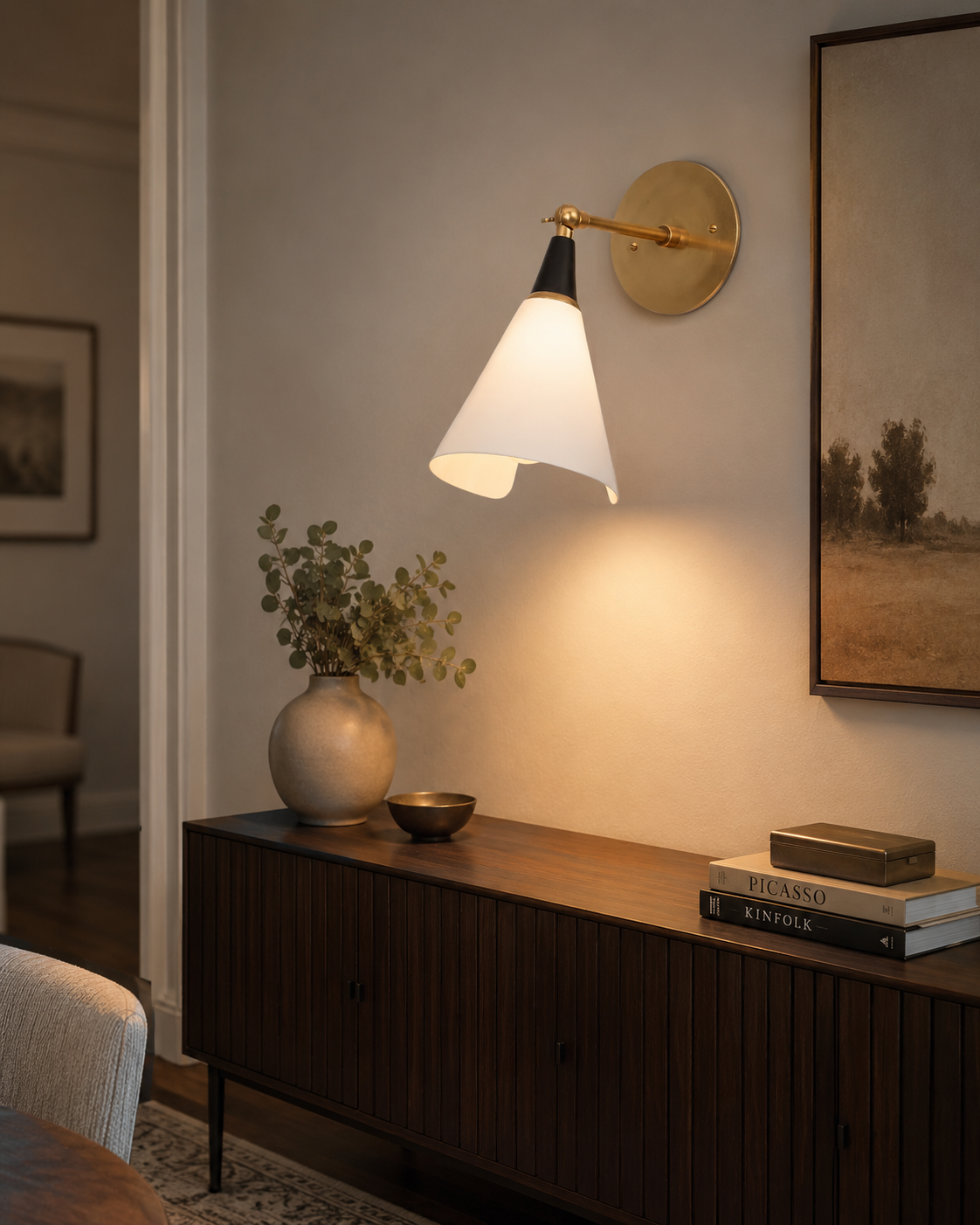 Milan Adjustable Cone Wall Sconce installed above a wooden sideboard in a modern living room, providing warm ambient lighting with its adjustable white cone shade and solid brass arm.