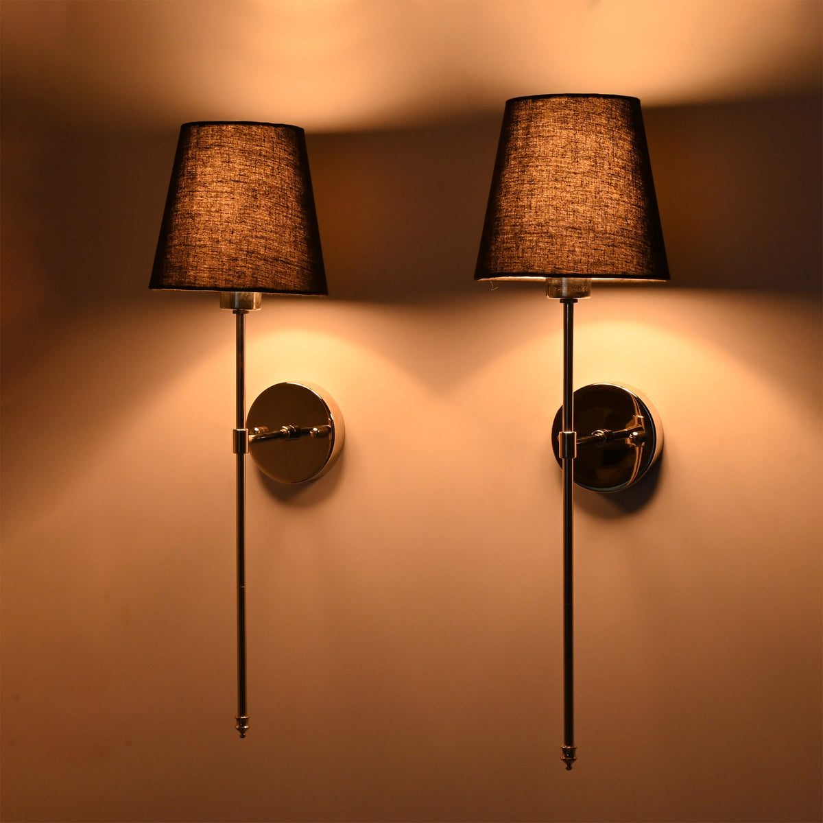 Front pair view of black shade chrome rod sconces glowing warmly with polished round wall plates.