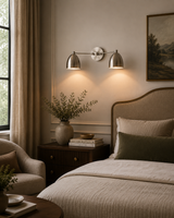 A modern two-light wall sconce with dome-shaped silver metal shades mounted on a beige wall above a bed in a styled bedroom setting.