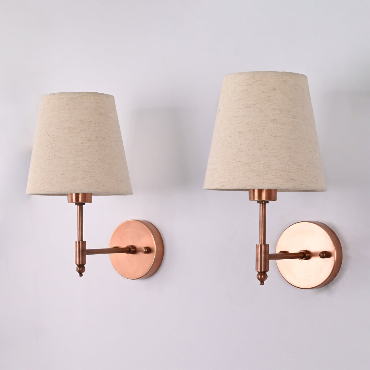 Pair of Milan copper antique brass wall sconces with natural linen tapered fabric shades displayed at an angle, highlighting the elegant wall arm design and handcrafted brass finish.
