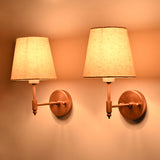 Pair of illuminated Milan copper antique brass wall sconces with natural linen shades viewed from an angle, showcasing the warm lighting and elegant handcrafted brass construction.