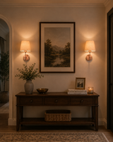 Two Milan copper antique brass wall sconces with natural linen fabric shades installed on either side of framed artwork above a wooden console table, creating a warm and elegant entryway with soft ambient lighting.