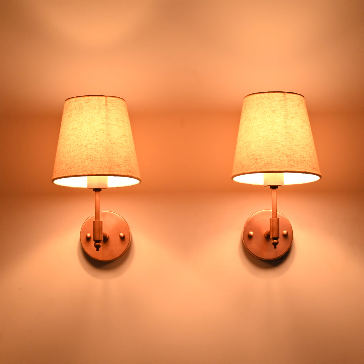 Front view of two Milan copper antique solid brass wall sconces with natural linen tapered shades illuminated to create soft, warm ambient lighting for modern and transitional interiors.