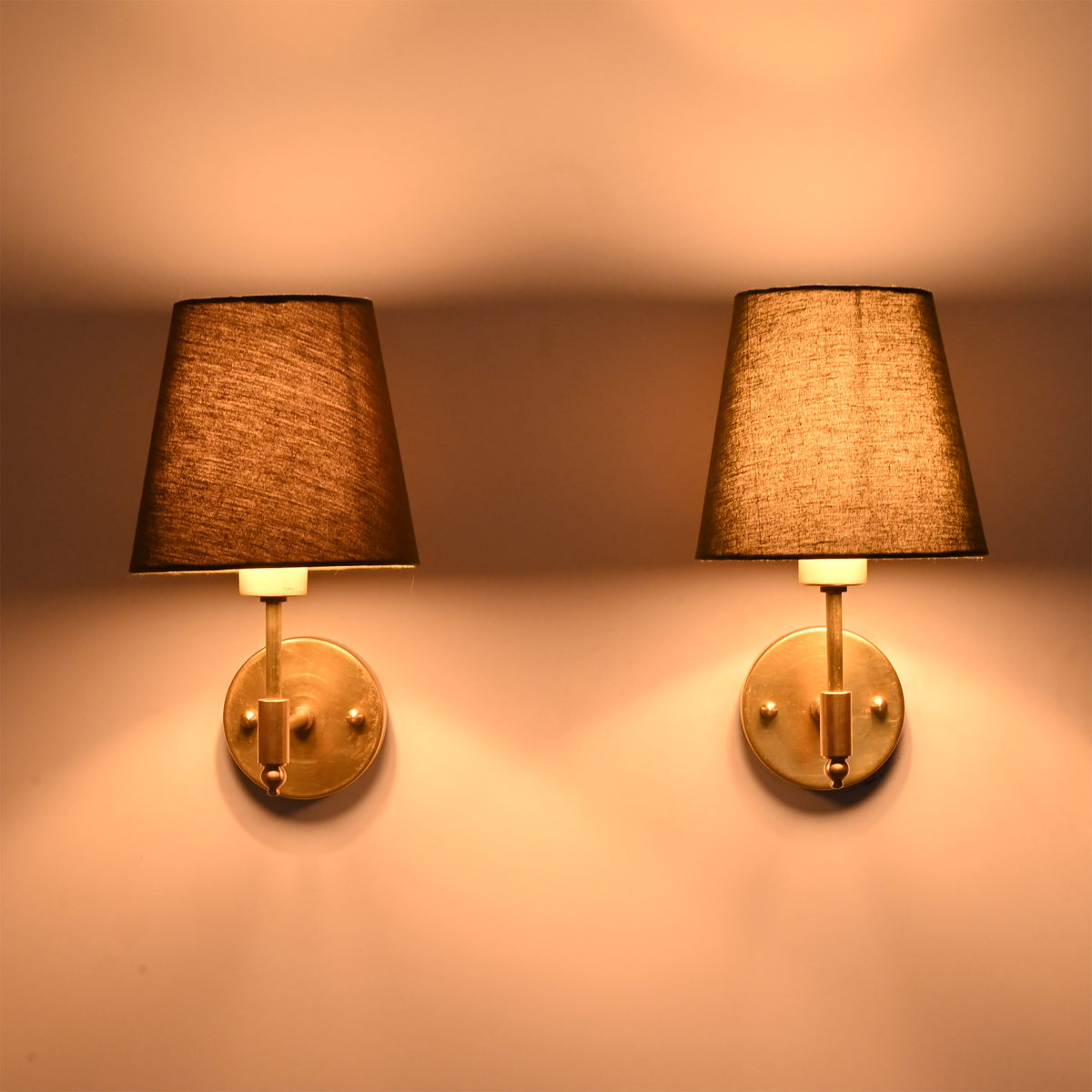 Illuminated Milan brushed brass wall sconces with olive green fabric shades creating a warm ambient glow against a neutral wall.