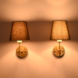 Illuminated Milan brushed brass wall sconces with olive green fabric shades creating a warm ambient glow against a neutral wall.