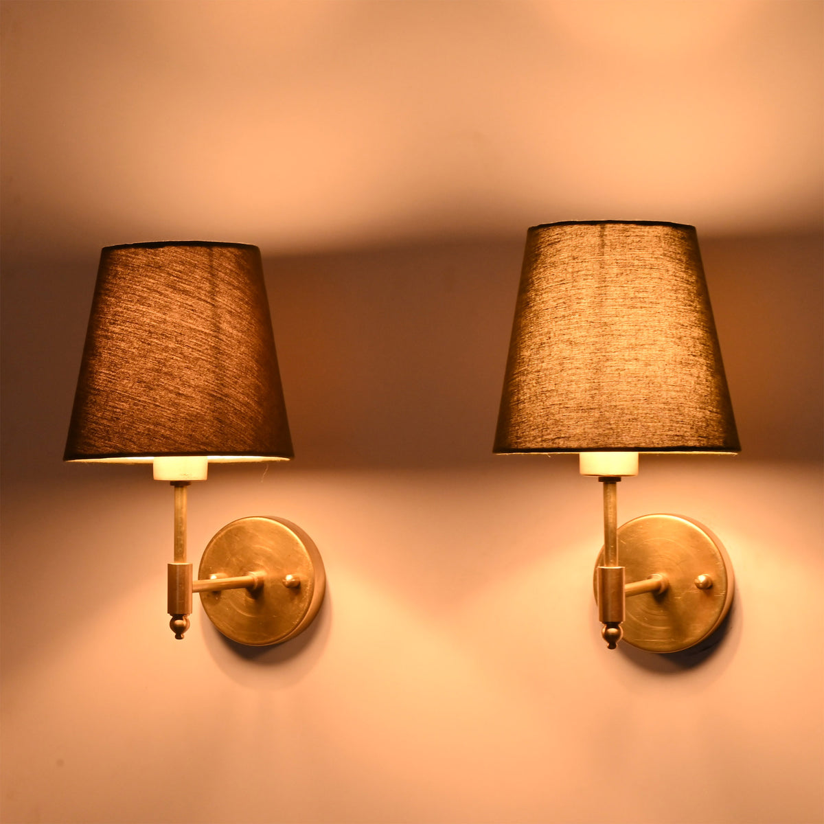 Side angle view of illuminated Milan brushed brass wall sconces with olive green tapered fabric shades showcasing warm ambient lighting.