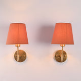 Front view of Milan Peach Shade Antique Brass Short Wall Sconce featuring a soft peach tapered fabric shade and handcrafted antique brass backplate.