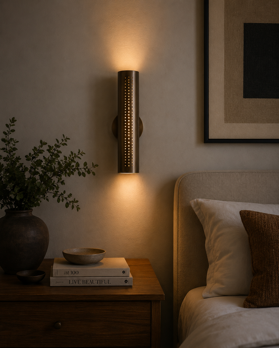 Milan perforated brass cylinder wall sconce illuminated beside a bed with warm ambient up and down lighting.
