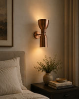 Milan sculpted hourglass 2-light copper wall sconce illuminated beside a bed in a warm modern bedroom interior.