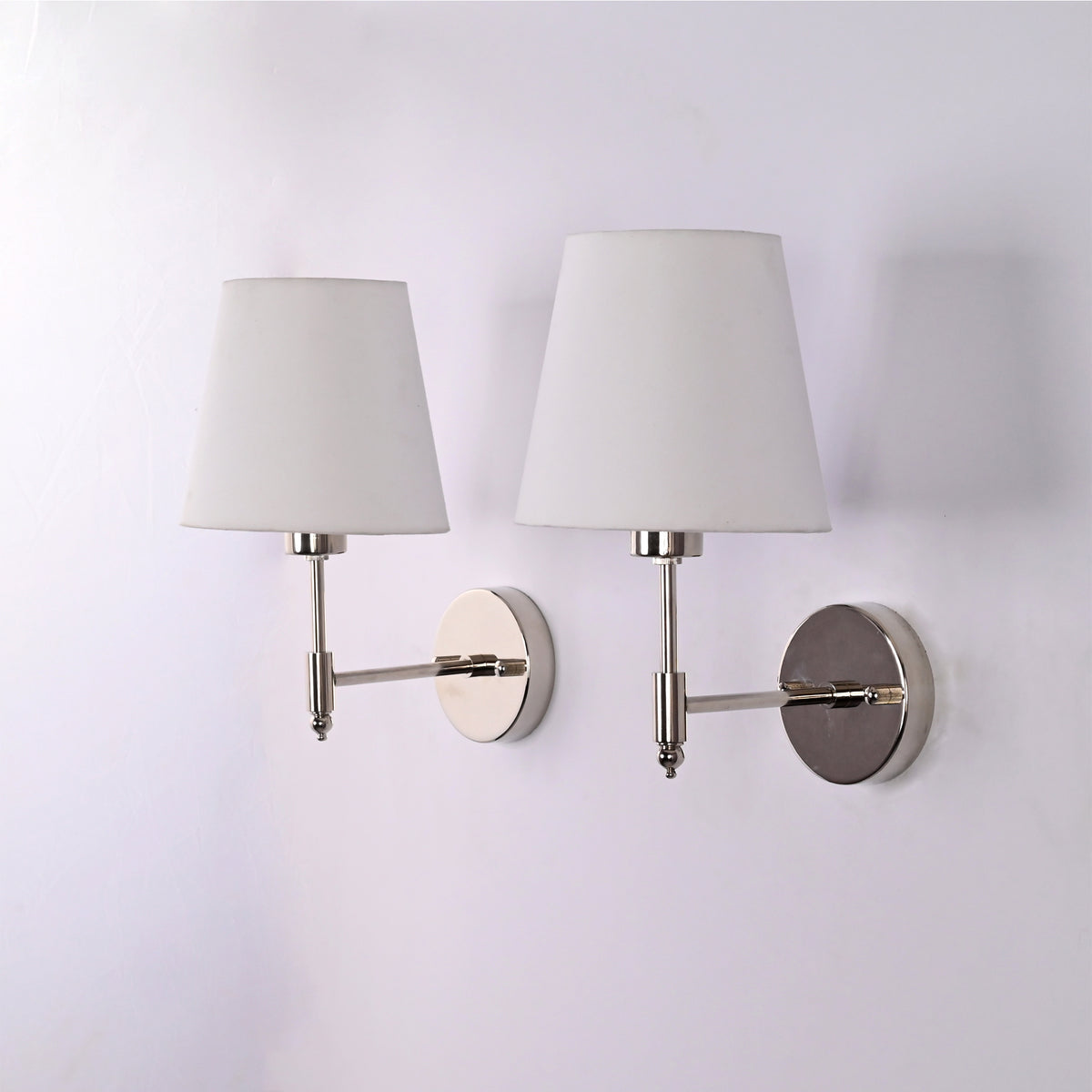 Angled view of two Milan polished chrome wall sconces featuring white tapered fabric shades and solid brass construction for contemporary interiors.