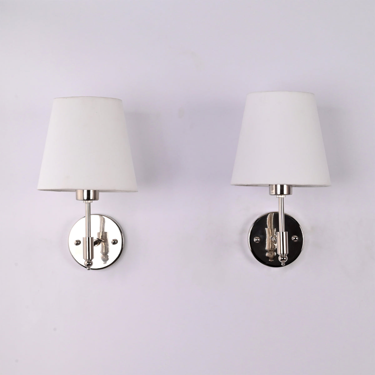 Pair of Milan polished chrome wall sconces with white tapered fabric shades mounted on a white wall, modern solid brass hardwired lighting fixtures.