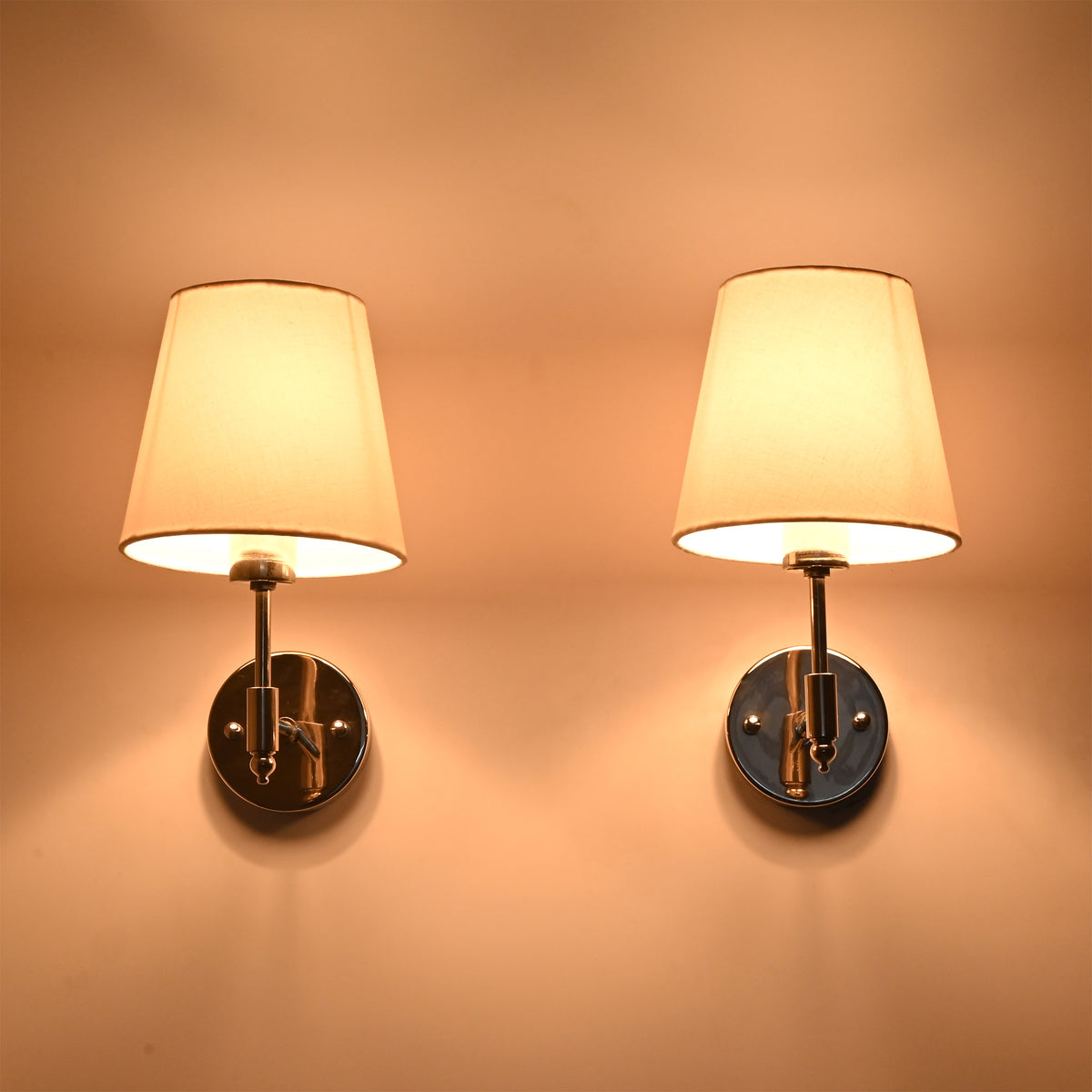 Pair of Milan polished chrome wall sconces with illuminated white fabric shades creating a soft warm ambient glow on the wall.
