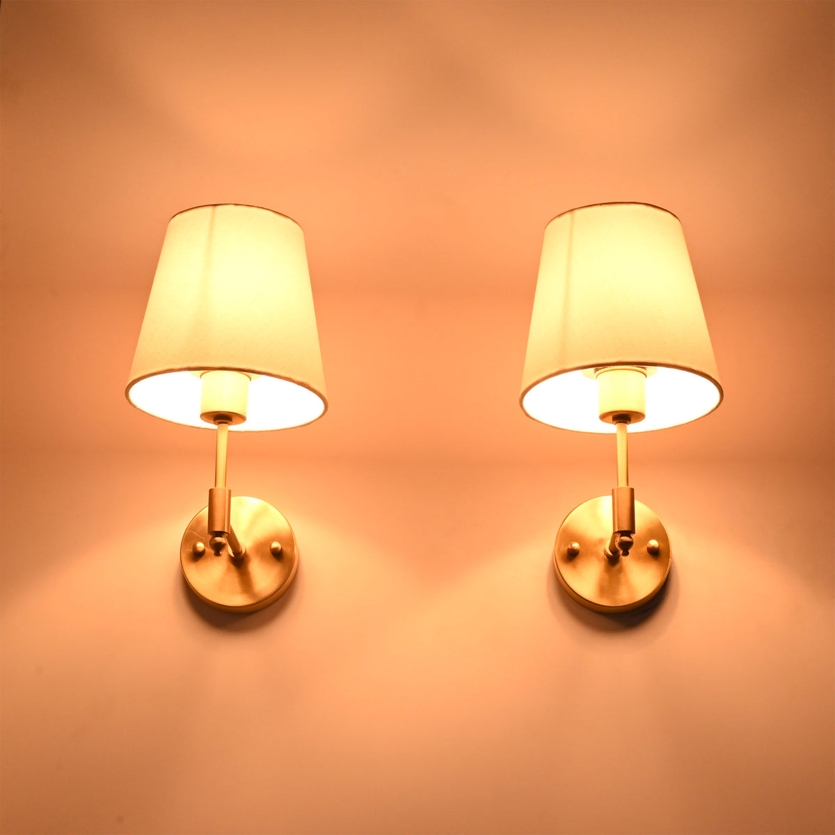 Illuminated Milan brushed brass wall sconces with white fabric shades casting a warm ambient glow against a neutral wall.