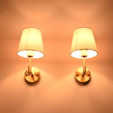 Illuminated Milan brushed brass wall sconces with white fabric shades casting a warm ambient glow against a neutral wall.
