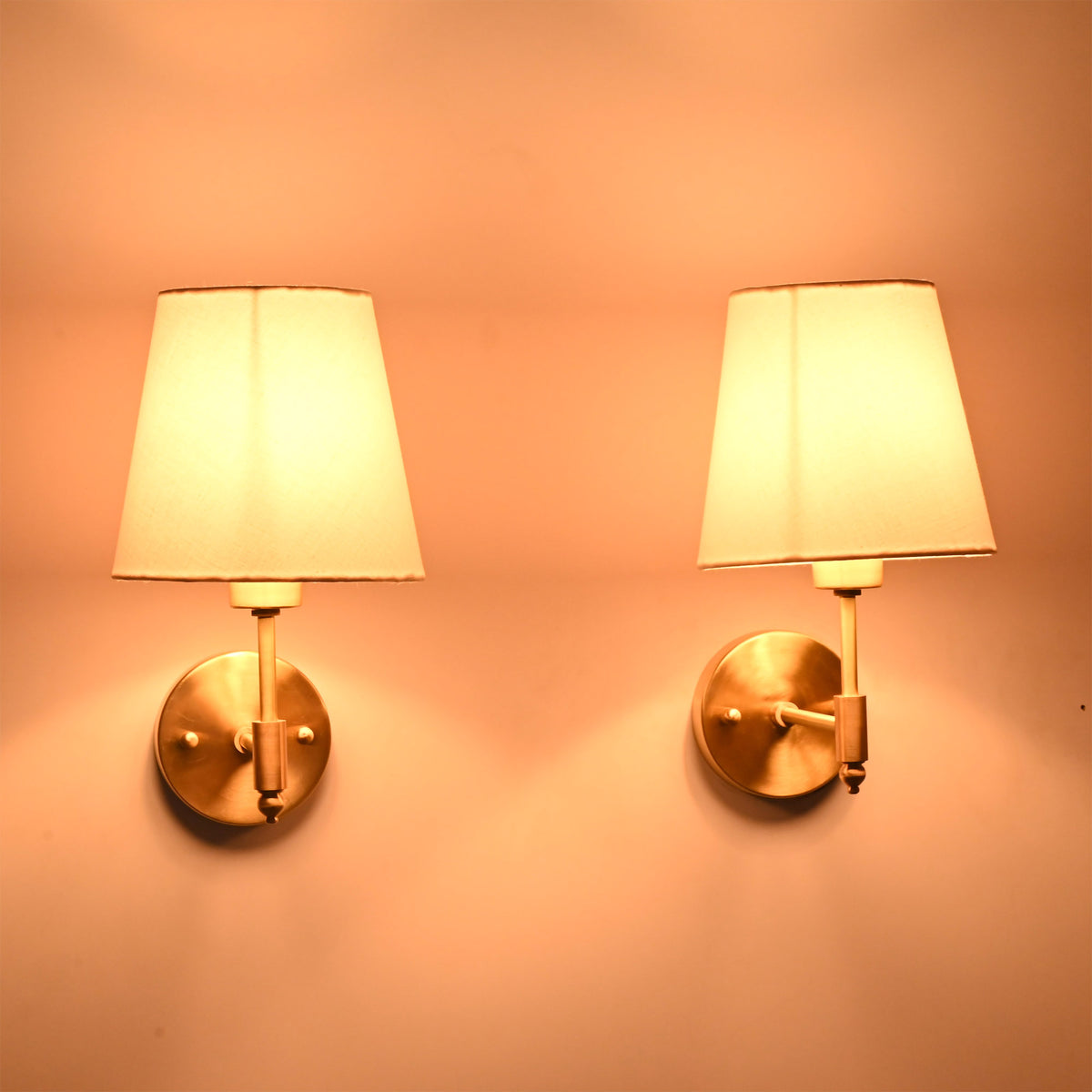 Perspective view of illuminated Milan brushed brass wall sconces with white fabric shades highlighting the handcrafted brass finish and soft lighting.