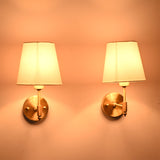 Perspective view of illuminated Milan brushed brass wall sconces with white fabric shades highlighting the handcrafted brass finish and soft lighting.