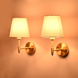 Side angle view of illuminated Milan brushed brass wall sconces with white tapered fabric shades showcasing warm ambient lighting.