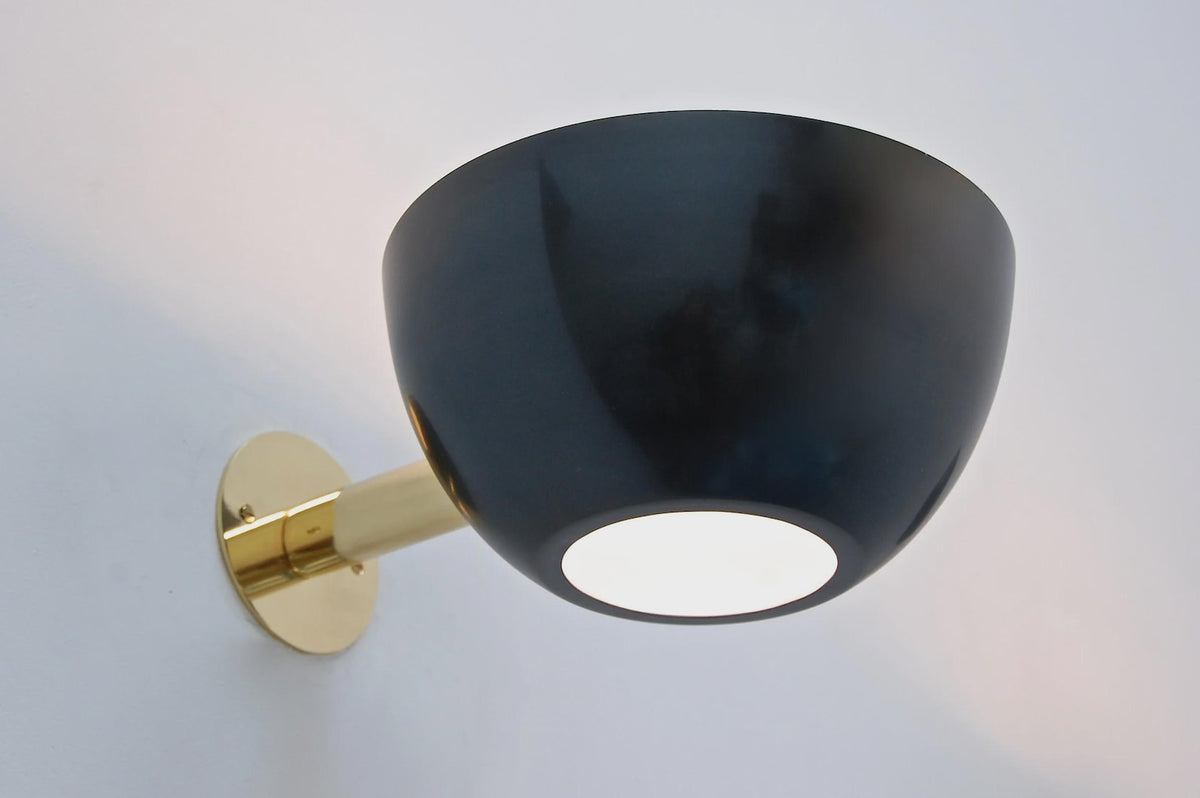 Angled view of a black bowl wall sconce with brass backplate, illuminated with warm light against a pale wall.