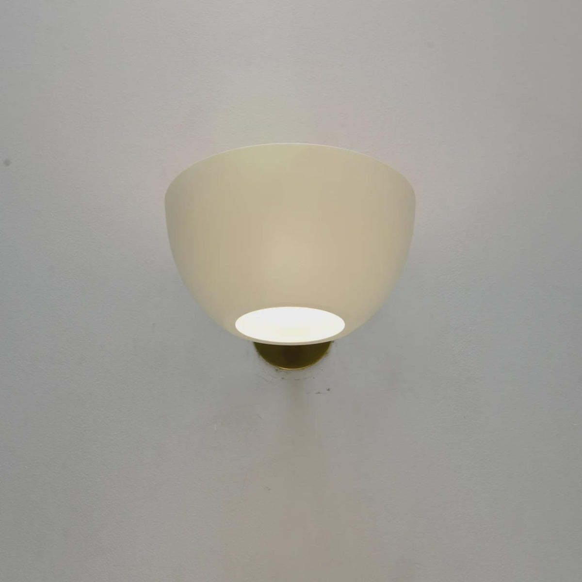 Centered white bowl wall sconce on a gray wall with warm diffused light and a brass mount visible beneath the shade.