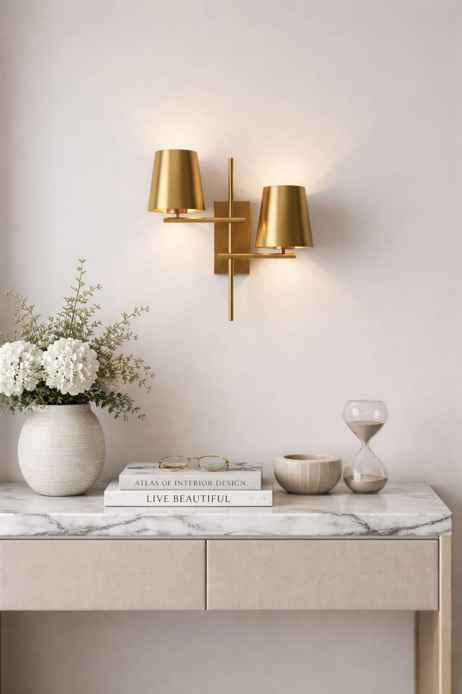 Modern 2-light brass wall sconce with dual tapered shades mounted above a marble-top console table with decor accents.
