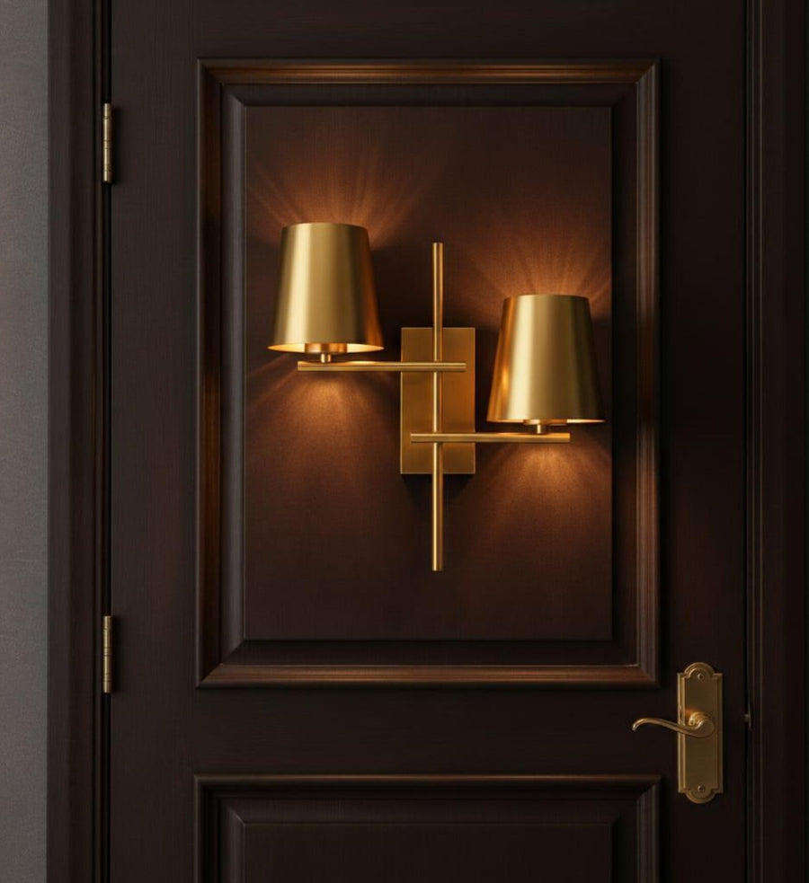 Modern 2-light brass wall sconce with dual tapered shades mounted above a marble-top console table with decor accents.