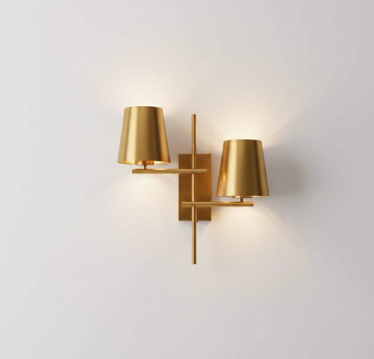Front view of a geometric brass wall sconce with two tapered shades and warm ambient light on a white wall.