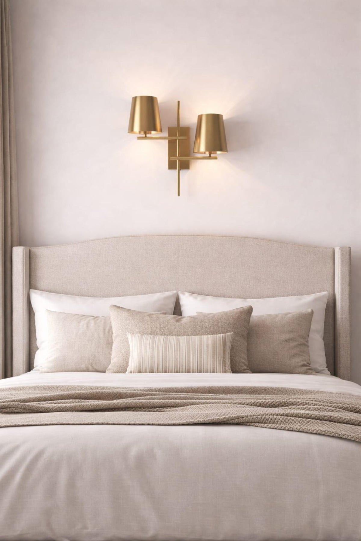 Brass 2-light wall sconce with twin tapered shades mounted above a bed in a soft neutral bedroom setting.