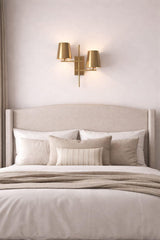 Brass 2-light wall sconce with twin tapered shades mounted above a bed in a soft neutral bedroom setting.