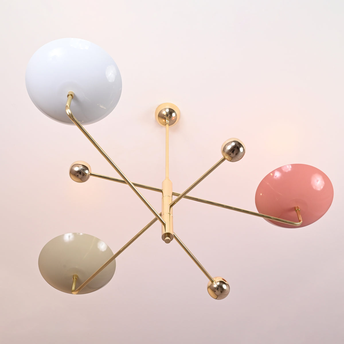 Modern three-arm brass chandelier viewed from below, featuring white, pink, and green painted shades with polished brass accents.