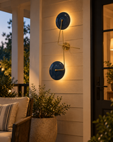 Nashville navy blue and brass wall sconce illuminating a covered porch at twilight beside a dark entry door, wicker seating, and greenery.