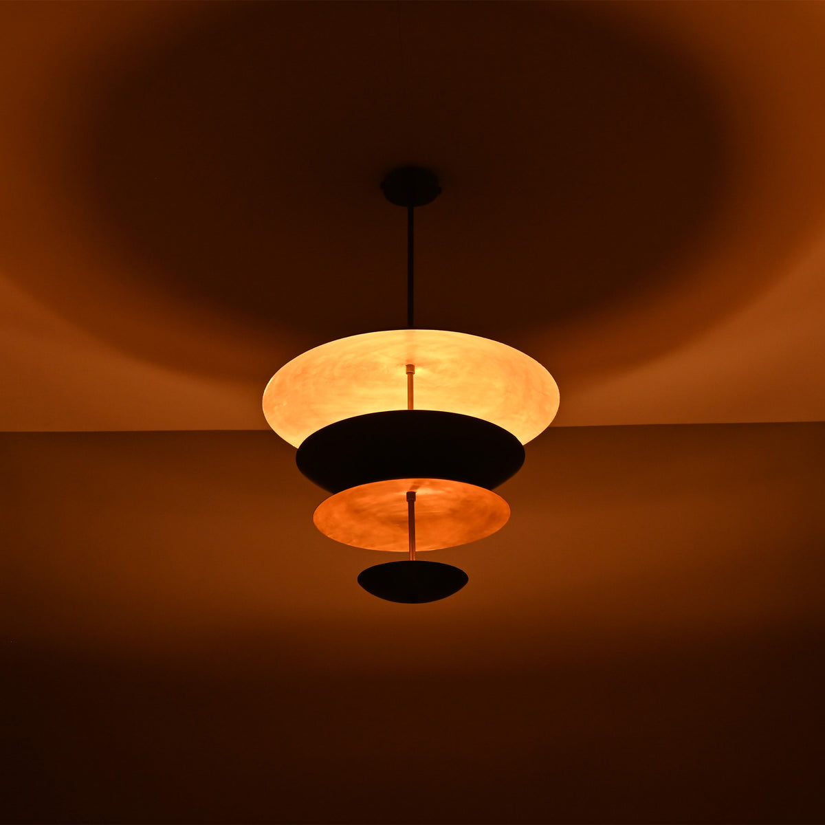 Noir Tiered Disc Chandelier glowing warmly in low light with dramatic ceiling and shade illumination.