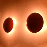 Two handmade red disc brass sconces on wall, illuminated with warm circular backlit glow for dramatic effect