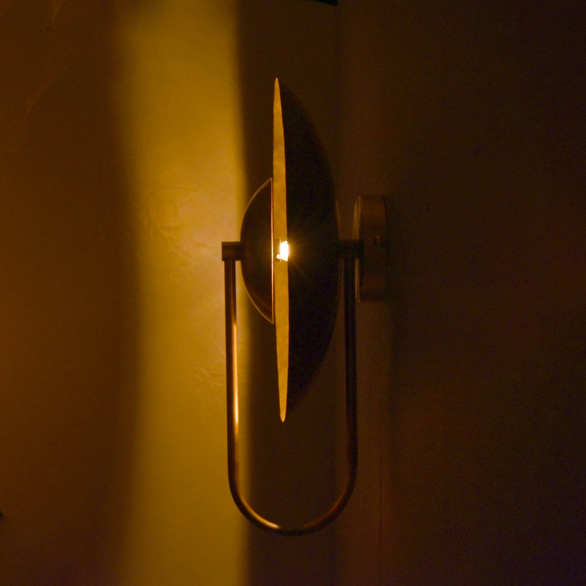 Slim side profile view of brass wall sconce illuminated in low light, highlighting circular glow effect