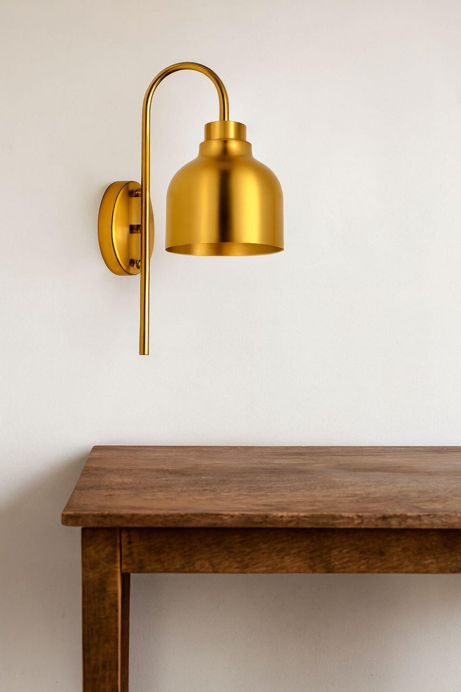 Modern brass wall sconce with dome bell shade mounted above a wooden console table, casting a warm ambient glow on a beige wall.
