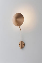 Modern brass disc wall sconce with round shade and curved arm casting a warm halo glow on a light wall.