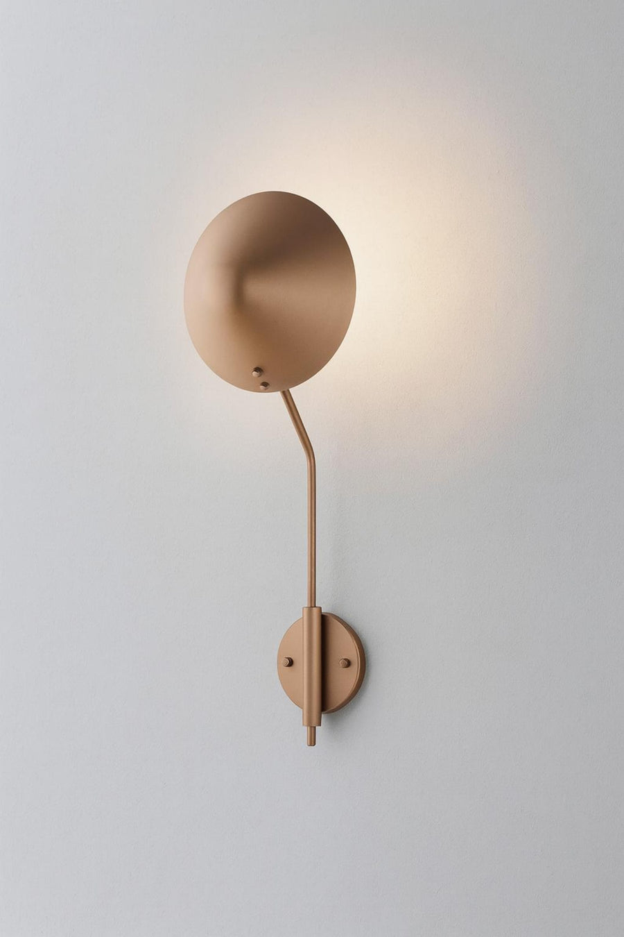 Modern brass disc wall sconce with round shade and curved arm casting a warm halo glow on a light wall.