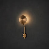 Brushed brass disc wall sconce illuminated on a dark wall, creating a circular halo glow and modern minimalist look.