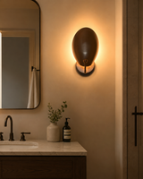 Sloane Modern Oval Shield Wall Sconce installed beside a modern bathroom vanity, creating a warm indirect ambient glow with its oversized handcrafted oval metal shield.