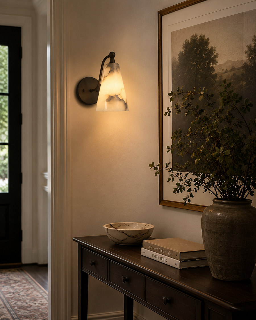 Sloane tapered alabaster wall sconce installed in a contemporary hallway interior, casting a warm ambient glow with its handcrafted natural alabaster cone shade and curved solid brass arm.