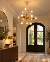 Tokyo 12-light brass-tone Sputnik chandelier illuminating an elegant entryway featuring arched black double doors, a wooden console table, decorative mirror, and warm ambient lighting.