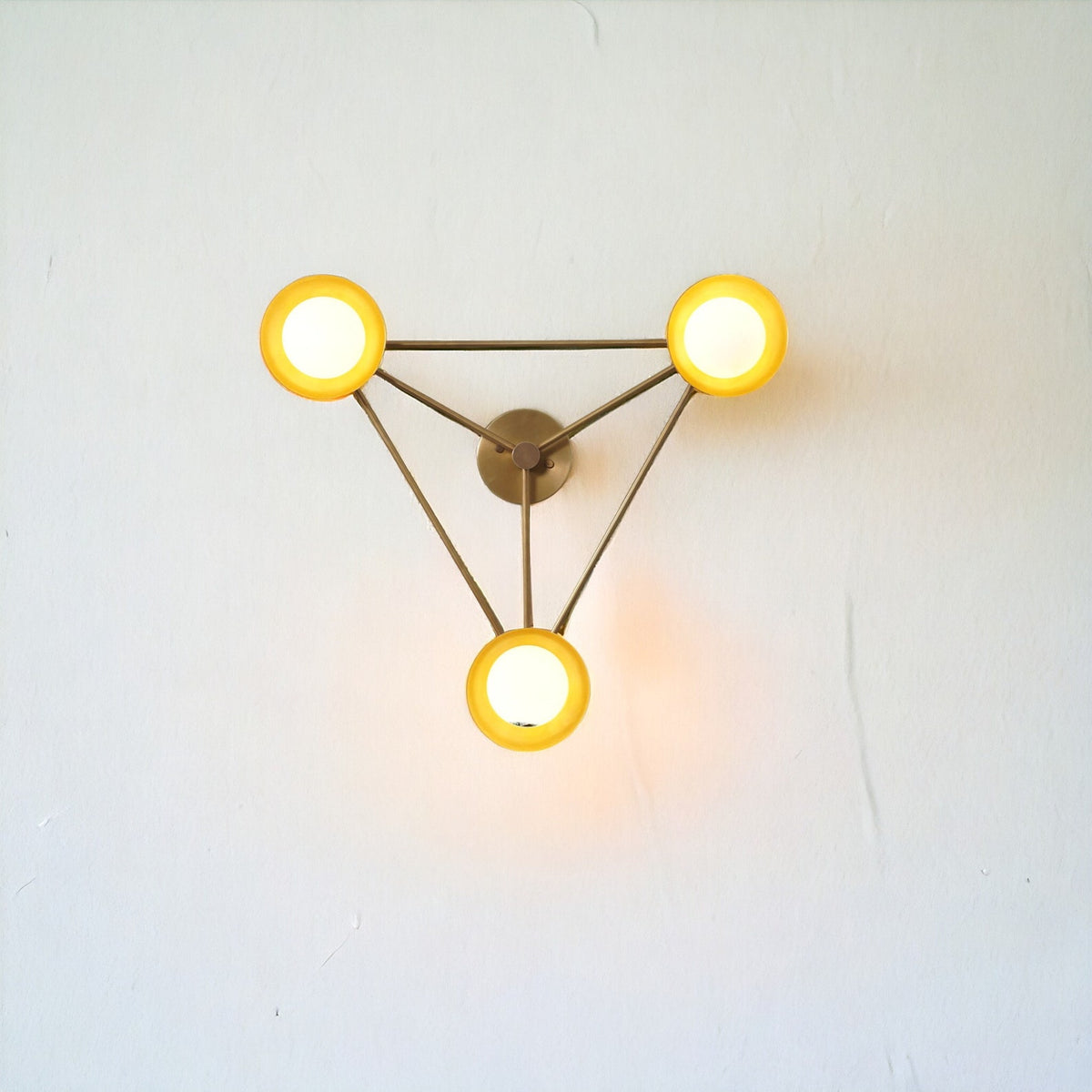 Tri-disc brass wall light mounted on a wall, displaying soft warm lighting and a geometric brass structure that complements modern interior decor.