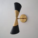 Angled right view of brass and black double cone wall sconce showing the round backplate and slim arm on a light wall.