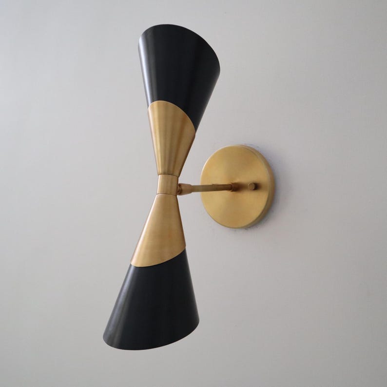 Angled left view of two tone brass and black double cone wall sconce with round brass backplate on a light wall.