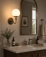 Verona modern globe glass wall sconce installed beside a bathroom mirror, providing warm ambient lighting above a marble vanity with antique brass fixtures.
